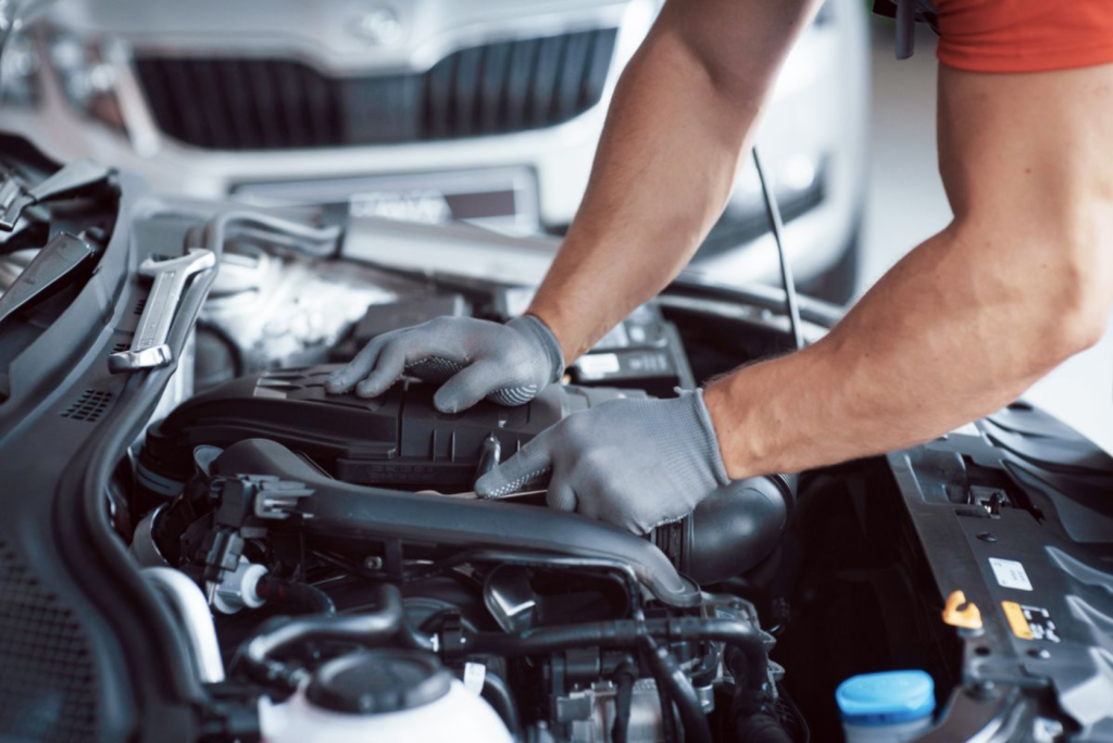 The image shows a person wearing gloves working on a car engine under the hood.
